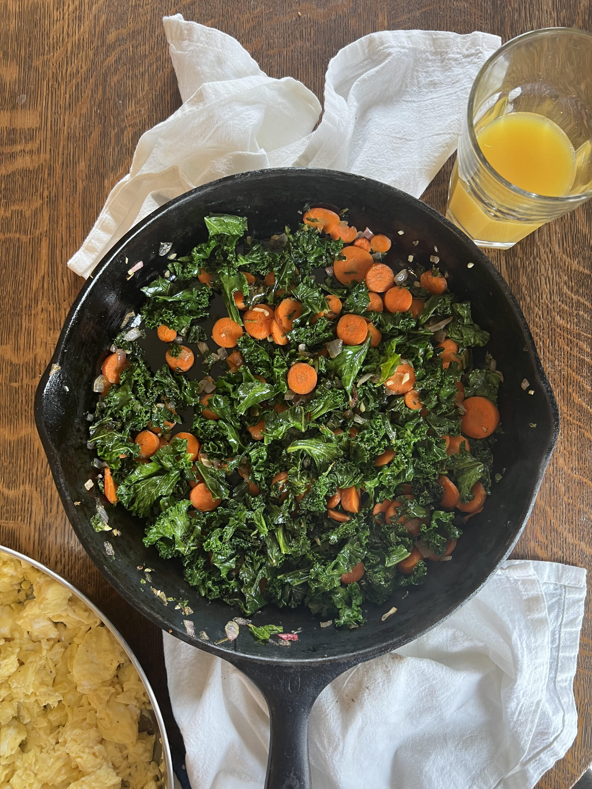 A pan with a veggie hash, a glass of orange juice, and a pan with scrambled eggs